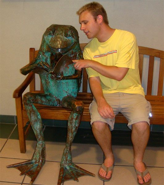 Russell gesturing to a statue of a frog reading on a bench.